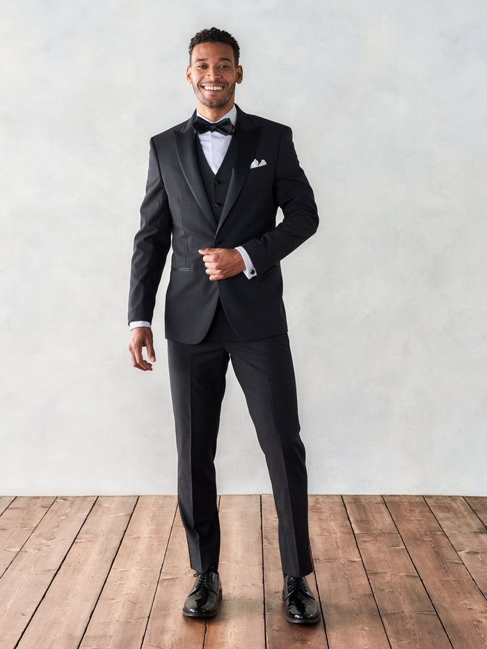 Smiling man in a black tuxedo with vest, bow tie, and white pocket square, on a wood floor.