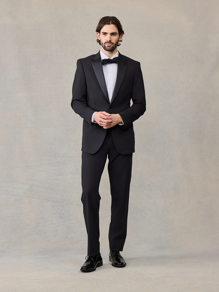 Man in a black tuxedo with satin lapels, hands clasped, black bow tie.