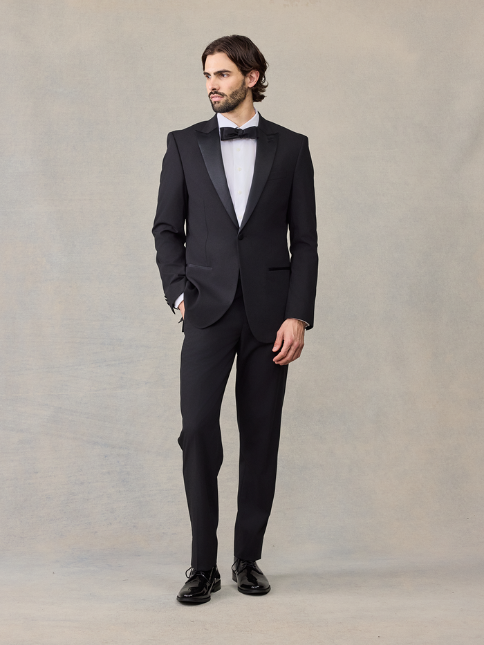 Man in a black tuxedo with a satin lapel jacket and black bow tie, full length.