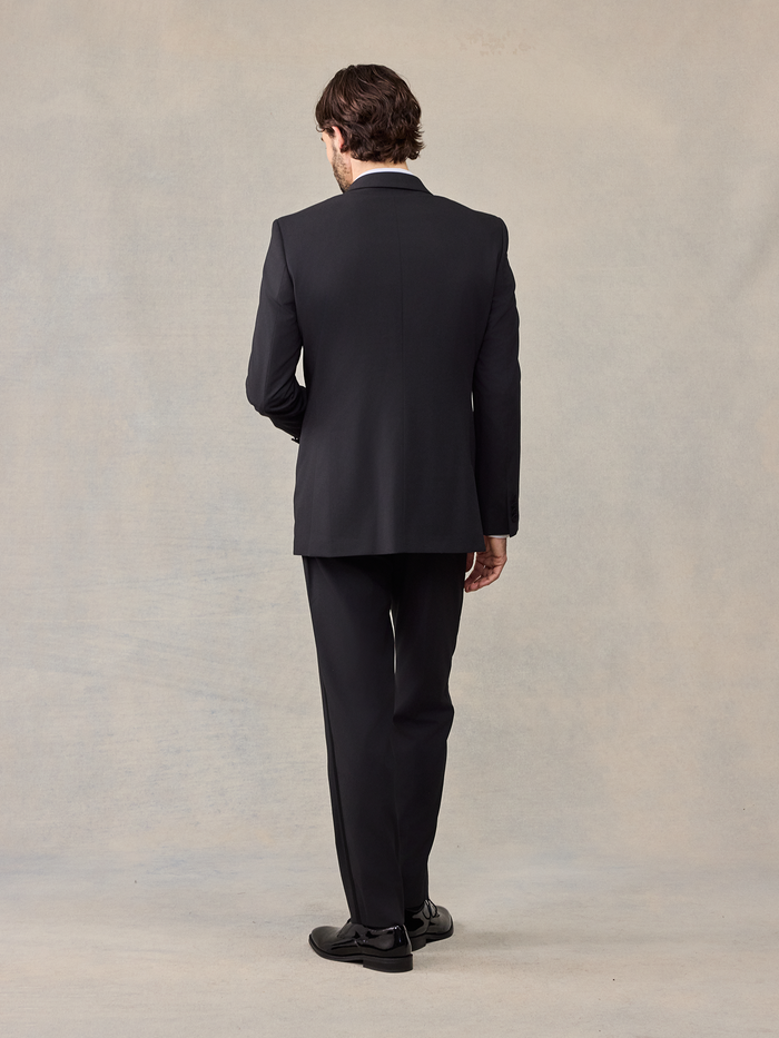 Back view of a man in a black tuxedo jacket and trousers, full length.