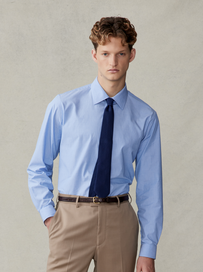 Man in a light blue dress shirt with a navy tie and beige trousers.