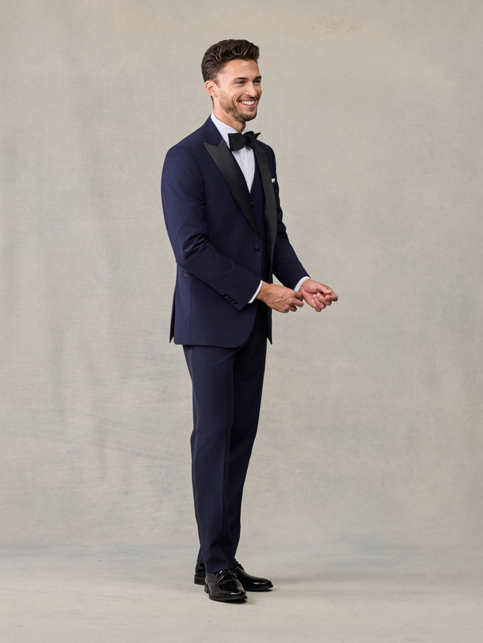 Side view of a smiling man in a navy tuxedo, hands clasped in front.