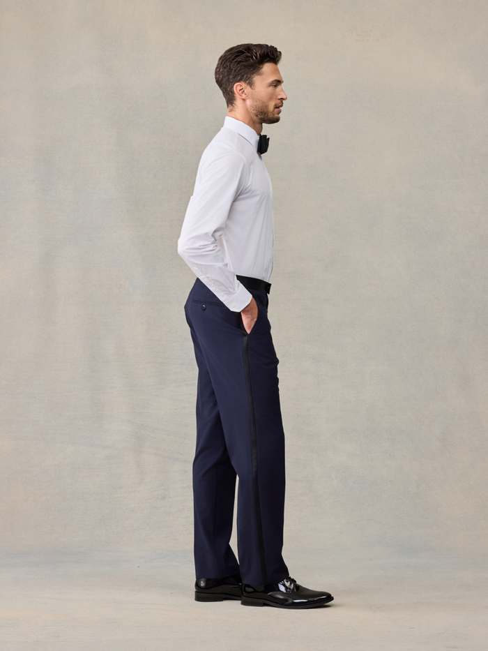 Side view of a man in a white tuxedo shirt and navy trousers, hand in pocket.