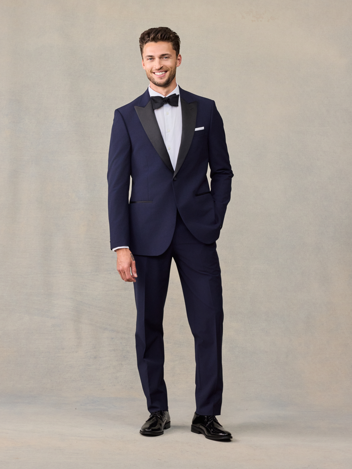 Smiling man in a navy tuxedo with black satin lapels and a black bow tie, full length.