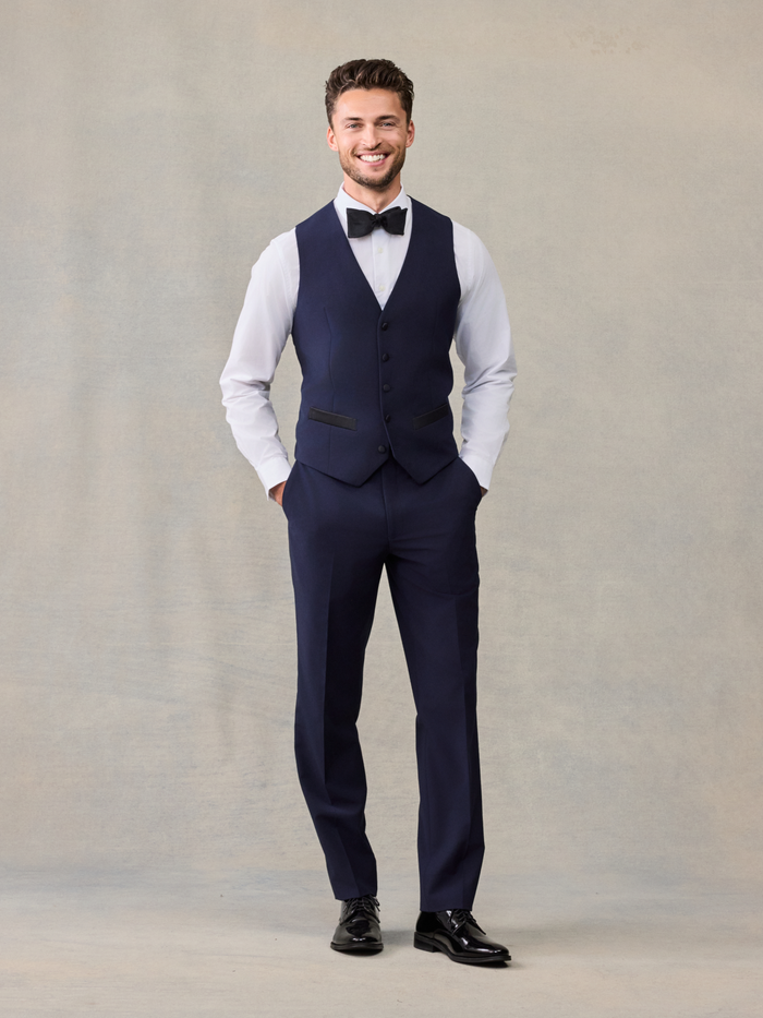 Smiling man in a navy tuxedo vest with a black bow tie, full length.