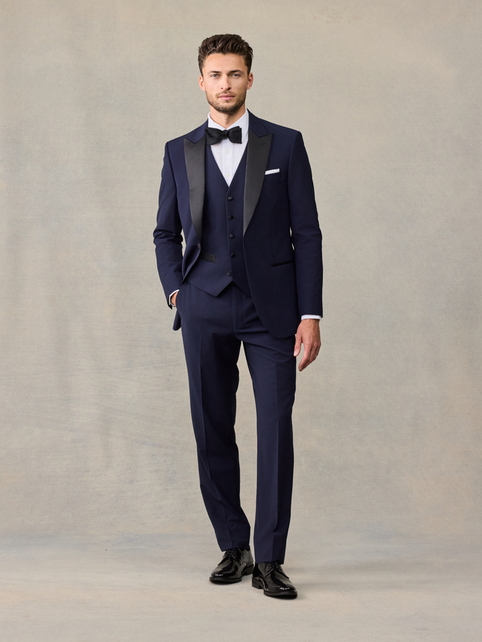 Man in a navy tuxedo with vest, black bow tie, and white pocket square, hand in pocket.