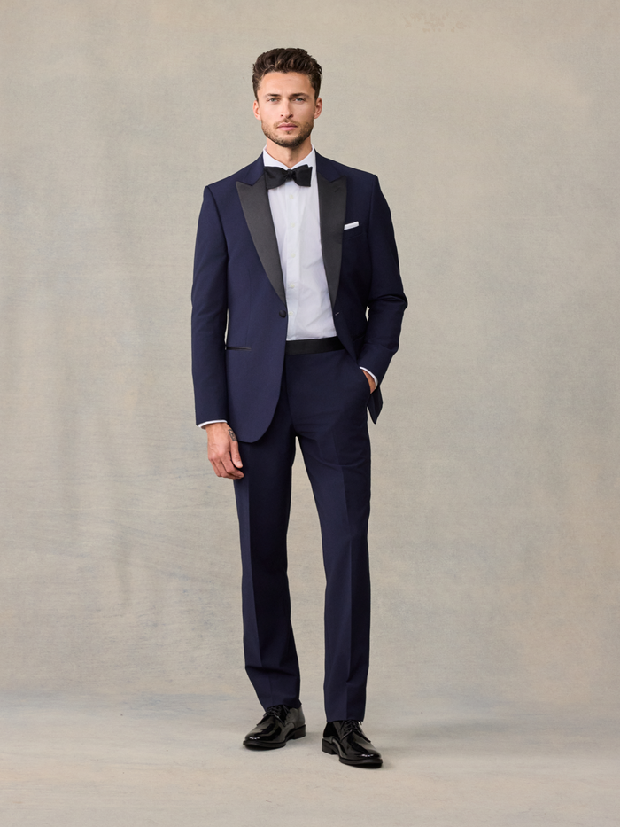 Man in a navy tuxedo with black satin lapels, hand in pocket, full length.