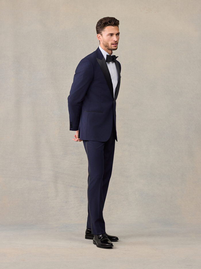 Side view of a man in a navy tuxedo jacket and trousers, hands behind his back.