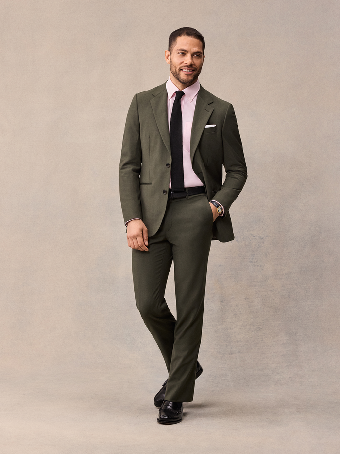 Man in an olive suit with a pink shirt and black tie, hand in pocket.