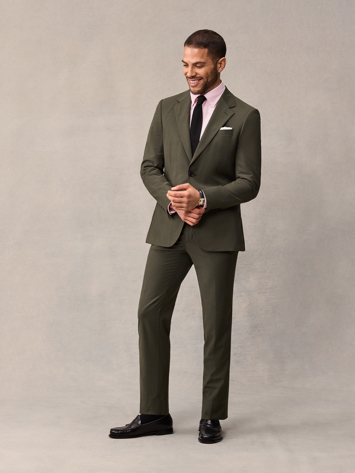 Man in an olive suit smiling and looking down, hands clasped together.