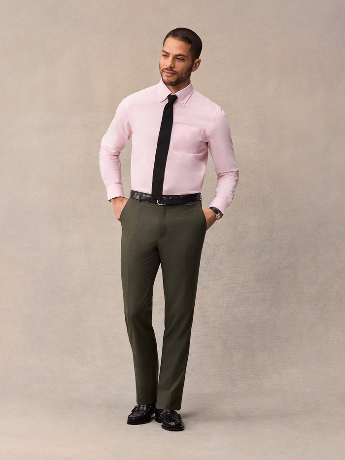 Man in a pink dress shirt and black tie with olive trousers, hand in pocket.