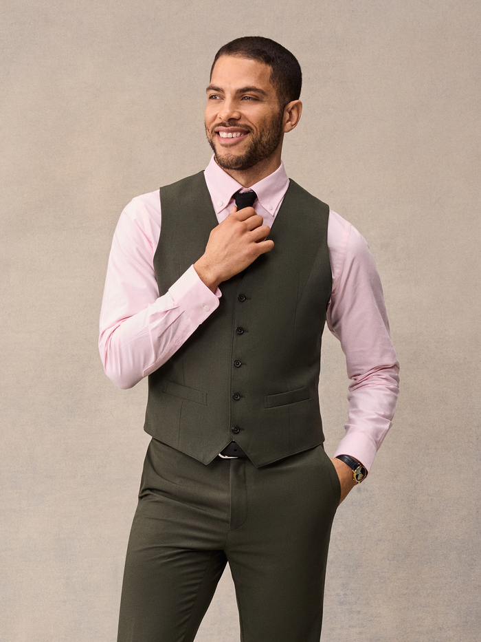 Man in an olive suit vest with a pink shirt, adjusting his black tie.