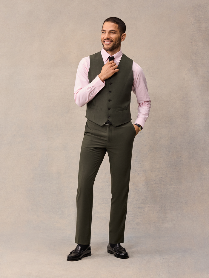 Man in an olive three-piece suit vest with a pink shirt, adjusting his tie.