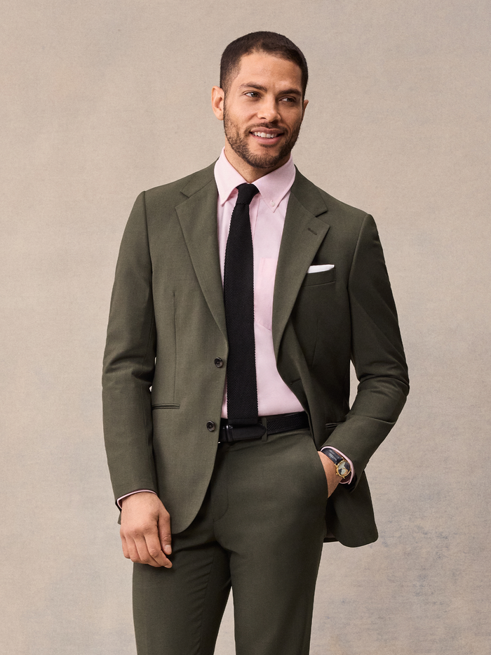 Man in an olive suit jacket with a pink shirt and black tie, smiling.