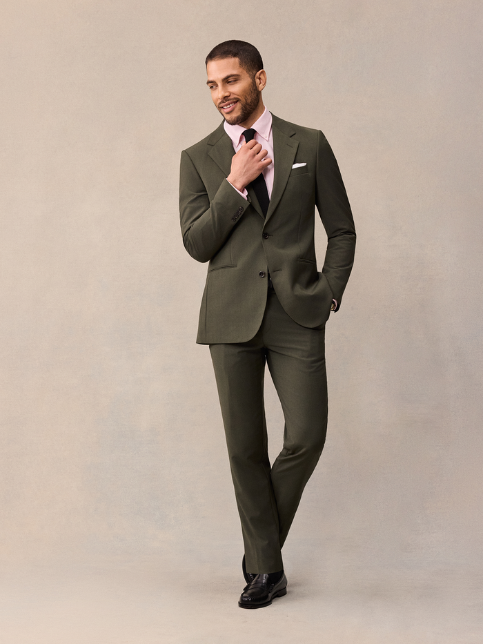 Man in an olive suit with a pink shirt and black tie, adjusting his collar.