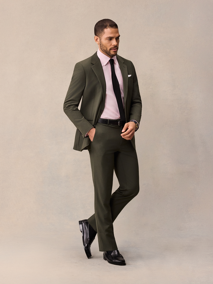 Man walking in an olive suit with a pink shirt and black knit tie.