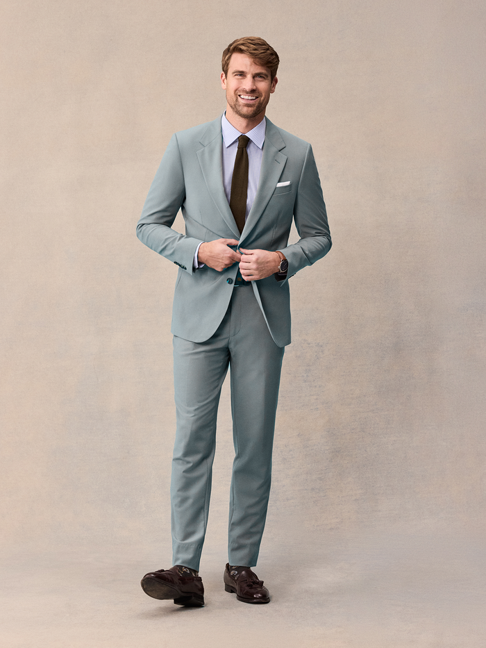 Man in a sage suit with a checked shirt, olive tie, and brown loafers.