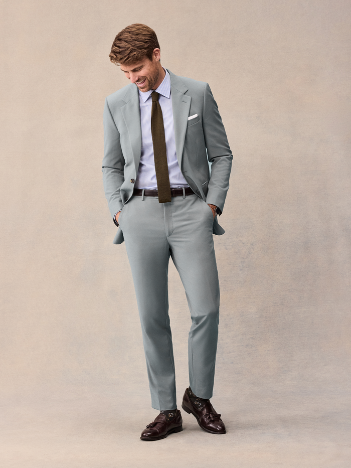 Man in a sage suit smiling and looking down, hands in pockets.