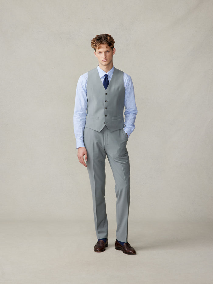 Man in a sage vest and trousers with a striped shirt and navy tie, full length.
