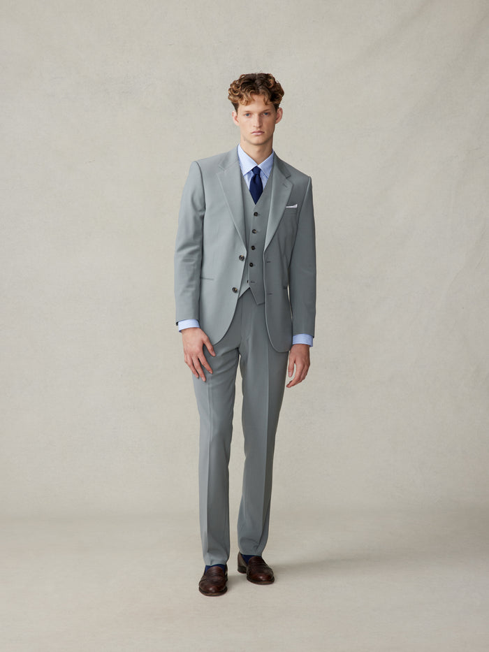 Man in a sage three-piece suit with vest, striped shirt, and navy tie, full length.