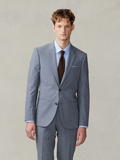 Man in a slate grey suit with light blue shirt, brown knit tie, and white pocket square, beige backdrop.