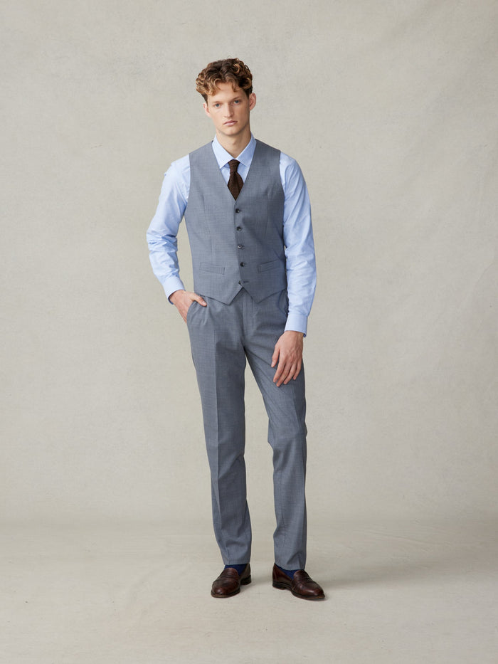 Man in a slate grey suit vest and trousers with a brown knit tie, full length.