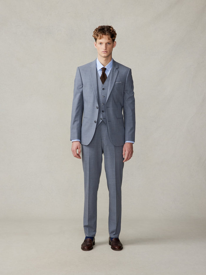 Man in a slate grey three-piece suit with a brown knit tie, standing.
