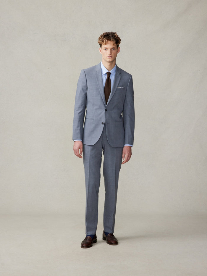 Man in a slate grey suit with a brown tie and brown shoes, full length.