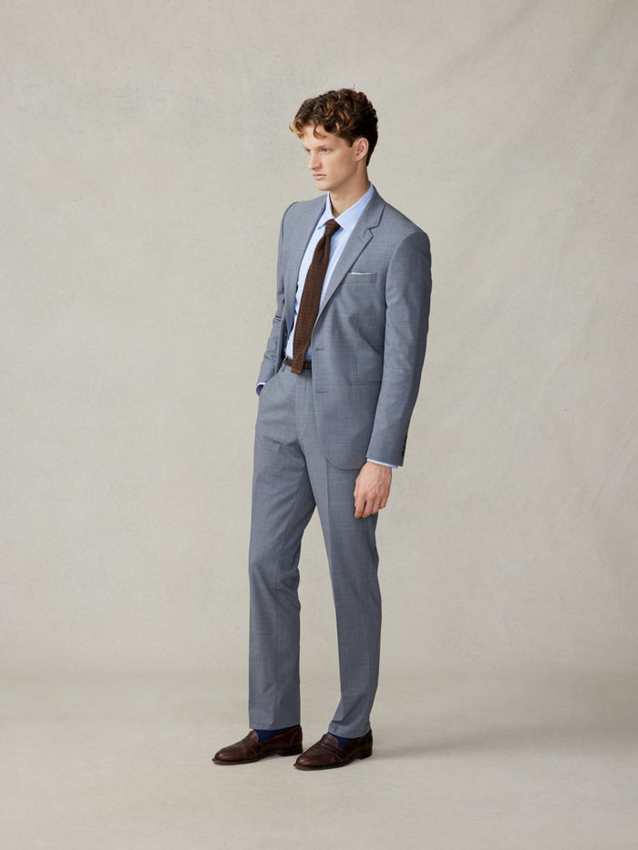 A man in a slate grey suit with blue shirt and brown tie, standing front view