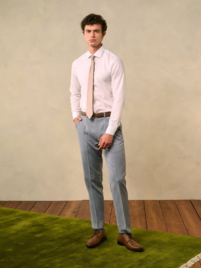 Man in a white shirt, tan tie, and sterling grey trousers, hand in pocket.