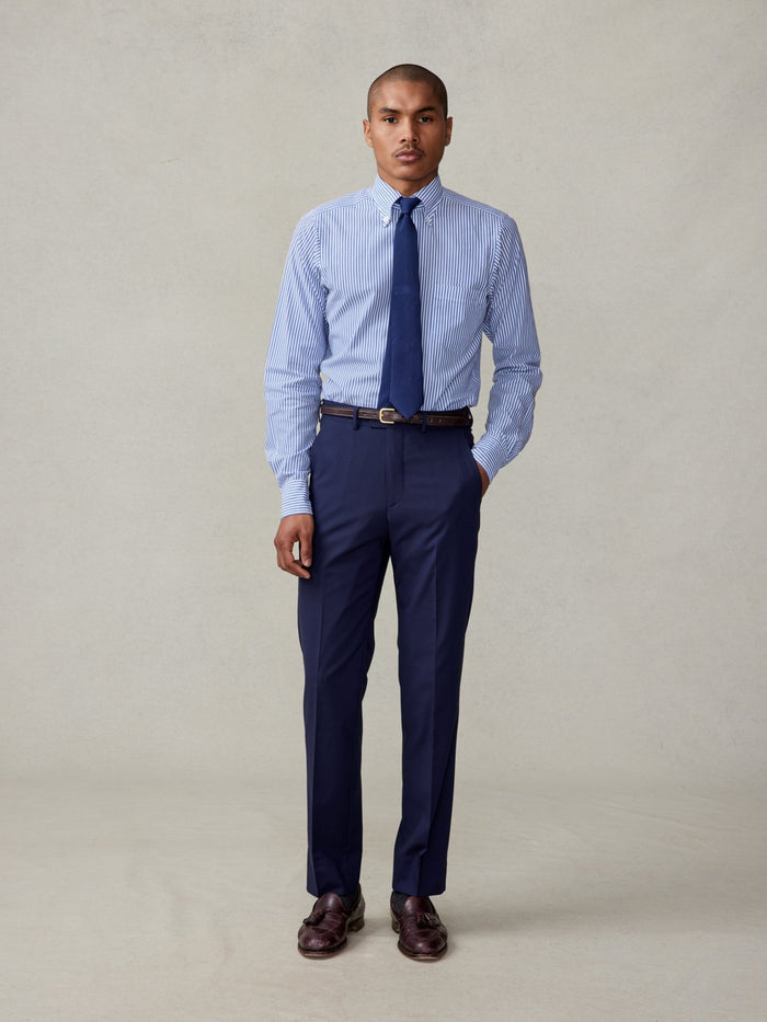 Man in navy suit trousers and a blue striped shirt with navy tie, front view, brown loafers, beige backdrop.