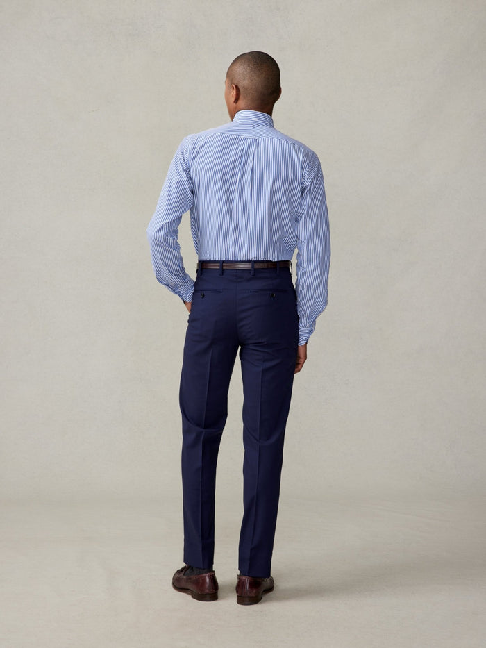 Man in navy suit trousers and a striped shirt, back view, full length, brown shoes, beige backdrop.