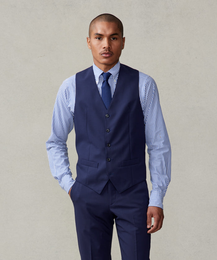 Man in a true navy suit vest with a striped shirt and navy tie, hand in pocket.