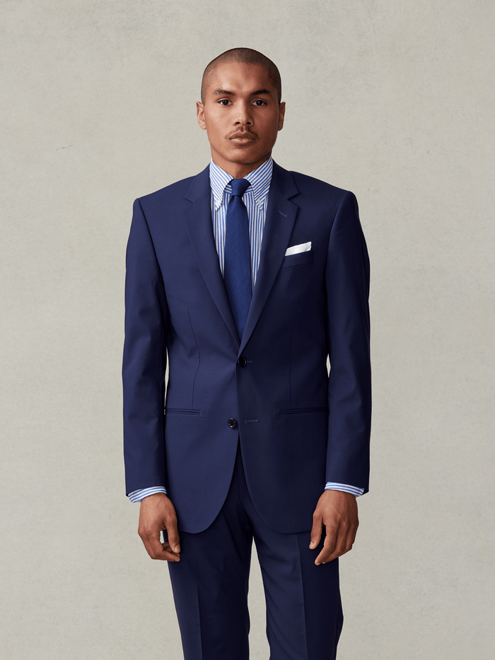 Man in a navy suit with blue striped shirt, navy tie, and white pocket square, beige backdrop.