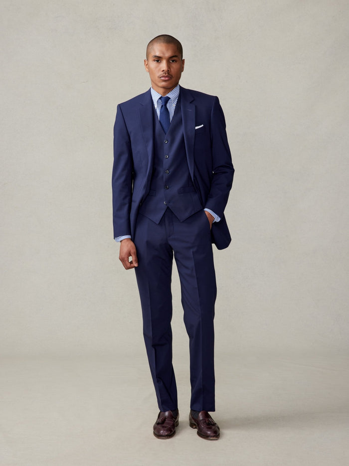 Man in a navy three-piece suit with vest, striped shirt, and navy tie, hand in pocket, beige backdrop.