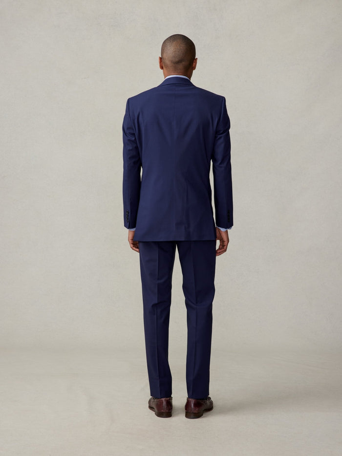 Man in a navy suit, back view, full length, striped shirt collar visible, beige backdrop.