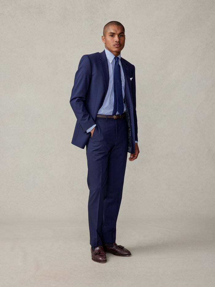 Man in a navy suit jacket over a striped shirt, side view, hand in pocket, full length, beige backdrop.