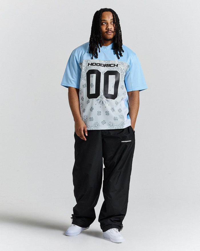 Person wearing a light blue t-shirt with a graphic design and text, black pants, and white shoes on a plain background.