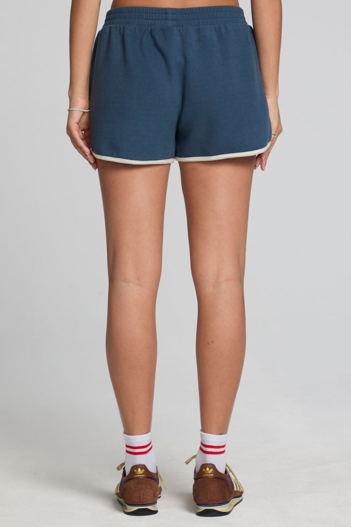 Back view of a woman wearing the Chaser  Eyla Dark Denim Shorts on a neutral background with brown sneakers and socks