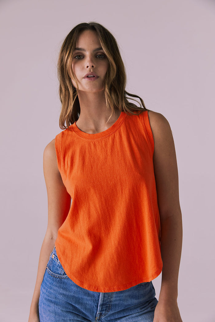 Front-facing studio image of the Chaser Flouncy Neon Flare Muscle Tank styled with denim shorts showing the bright neon orange hue and relaxed curved hem