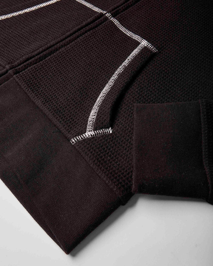 Close-up of a dark brown textured knit garment, showing a ribbed cuff, a waffle-knit panel, and contrasting white zigzag stitching on a light gray background.