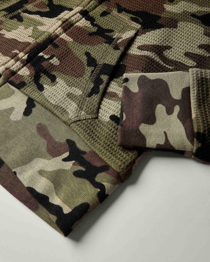 Close-up of a camouflage knit fabric garment, showing a waffle-knit texture with a pocket and folded cuff in shades of olive green, brown, beige, and black.