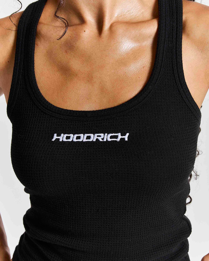 A close-up of a woman wearing a black waffle-knit tank top with the brand name 'HOODRICH' printed in white across the chest.