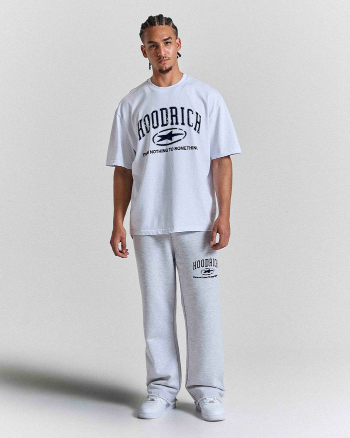 A man modeling a white short-sleeve t-shirt and light gray sweatpants set from the brand Hoodrich, featuring black collegiate-style graphic text and logos on both pieces, against a plain light gray studio background.