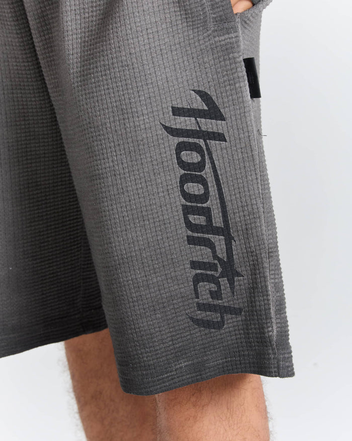 Close-up of a person wearing dark gray waffle-knit shorts with a large black vertical 'hoodrich' logo printed on the left leg, against a light gray background.