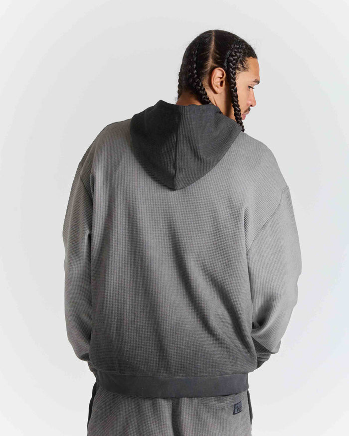 A man wearing a gray textured hooded sweatshirt and matching sweatpants, shown from the back against a light gray studio background.
