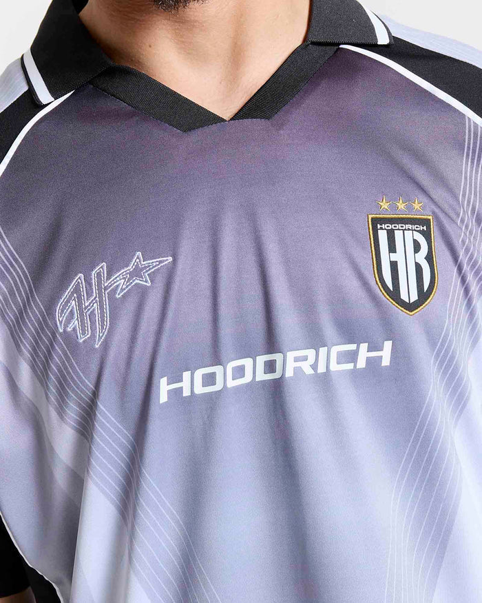 Close-up of a HOODRICH soccer jersey with a dark gray and light blue gradient, featuring the HOODRICH sponsor logo, the team crest, and a stylized star logo on the chest.