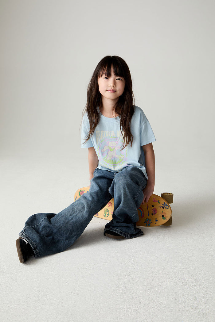 Image of a girl wearing the Chaser Grateful Dead In The Park Girls Tee on a neutral background sitting on a skateboard with one leg out