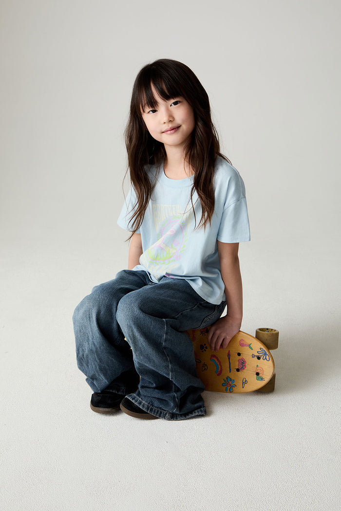 Image of a girl wearing the Chaser Grateful Dead In The Park Girls Tee on a neutral background sitting on a skateboard turned on its side