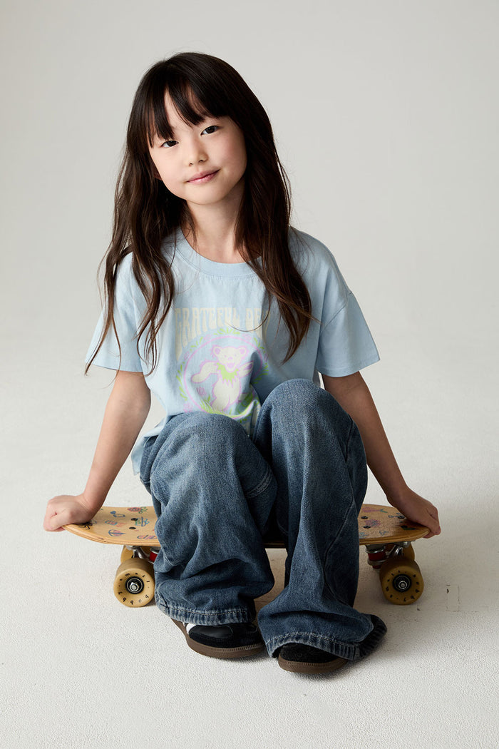 Image of a girl wearing the Chaser Grateful Dead In The Park Girls Tee on a neutral background sitting on a skateboard